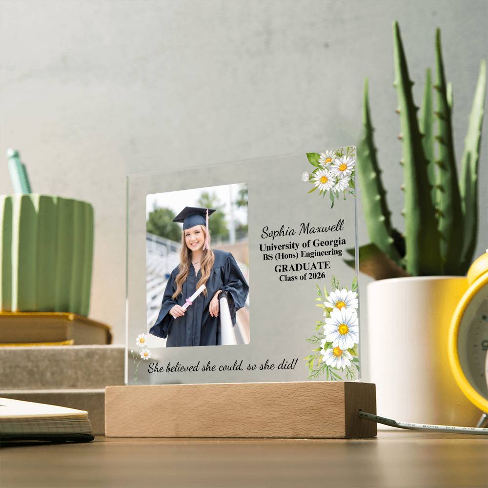 Graduation-themed decorative plaque with a photo of a graduate and text on a desk with plants and stationery.